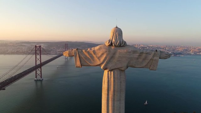 Aeria view monument Sanctuary of Christ the King. Drone flyby past near giant sculpture overlooking city of Lisbon Almada and famous 25th april bridge over river Tagus at sunset