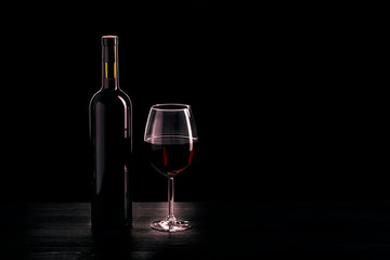 Bottle of red wine and a glass half filled with red wine, on a wooden black table, black background.