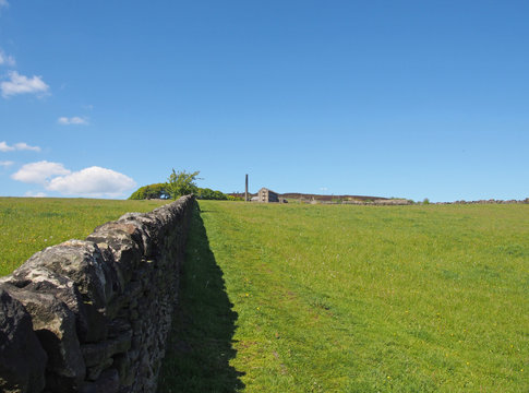 a long stone wall in perspective bordering a green grass meadow in bright spring sunlight with a historic abandoned mill and the village of old old town next to midgley moor in west yorkshire