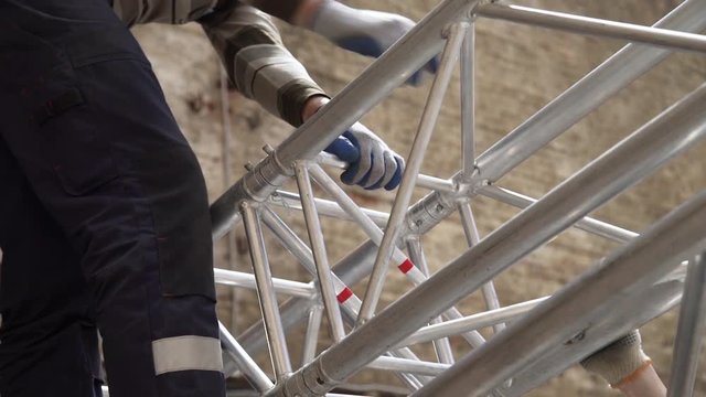 Man technical specialist is adjusting massive grey aluminium stage structure by hitting with hammer pins-connecters and fitting them together at daylight on clear sunny day.