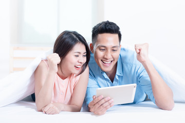 Asian young couple using a tablet while lying on the bed in the bedroom. High speed internet 5G service technology at home, Telecom networks designed concept.