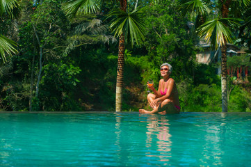 natural lifestyle portrait of attractive and happy middle aged Asian woman relaxed at tropical resort infinity simming pool with jungle background enjoying a drink relaxed