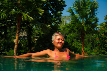 attractive and happy middle aged Asian woman relaxed at tropical resort infinity swimming pool with jungle background enjoying relaxed in luxury holidays trip