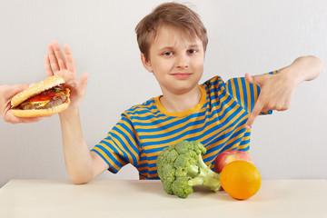 Young funny boy in a striped shirt at the table refuses hamburger in favor of fruit and vegetables...
