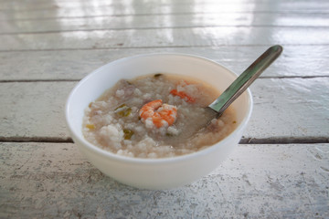 Rice congee mixed with shrimp and pork in white bowl and spoon on wooden table.