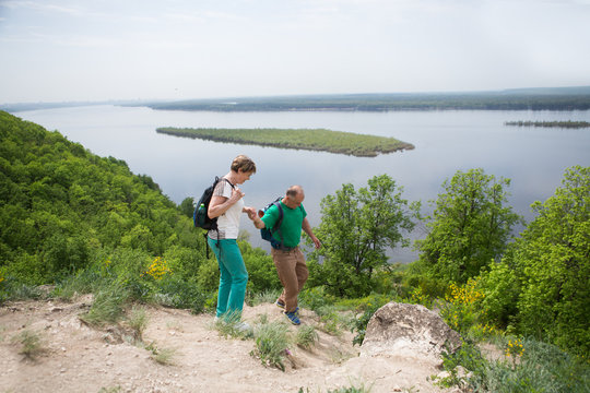 Elderly Couple With Backpacks Travels Around Mountains. Senior Couple Walking In Nature