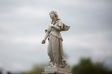 Statue of an angel holding a holy cross to it's chest. The angel is part of a gravestone. In the background cloud covered sky.