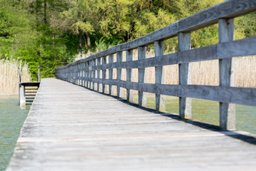 Obraz premium Wooden pier / footbridge at Lake Ammersee, photographed from the water towards the land. Wooden handrail on the left, trees and bushes in the background.