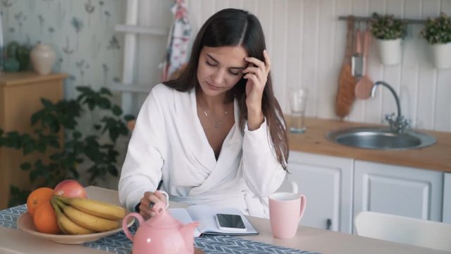 Young woman sits on kitchen, makes notes in notebook than looks away, slow motion