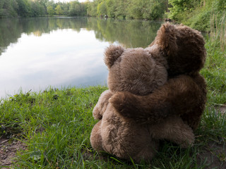 Zwei Teddybären sitzen Arm in Arm an einem See. Es ist ein romatischer Abend.