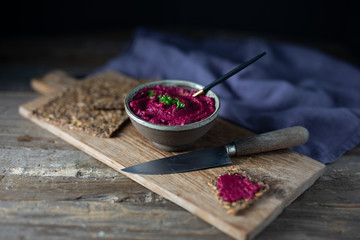 Focus on beet or beetroot hummus with parsley served with seed crackers on a rustic wooden board with a spoon, a rustic wooden knife and purple linen napkin. Side view copy space wooden background.