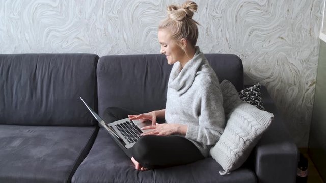 Cheerful young blond woman sitting on couch in living room and using laptop