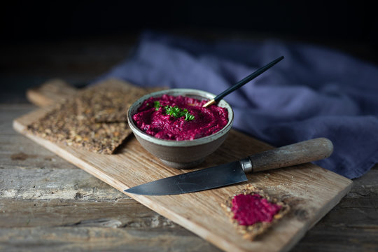 Focus on beet or beetroot hummus with parsley served with seed crackers on a rustic wooden board with a spoon, a rustic wooden knife and purple linen napkin. Side view copy space wooden background.