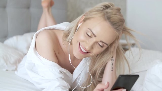 Beautiful, smiling blond woman lying in white bed and using a smartphone