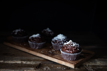 Chocolate cupcakes decorated with a chocolate cream frosting with coconut powder on top and chocolate shavings on a rustic wooden board on rustic wooden background.