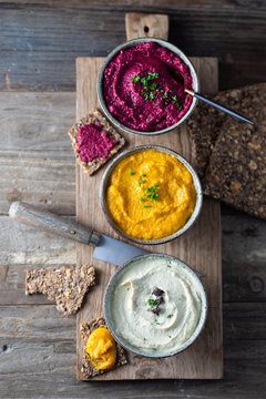 Three hummus dips, beet hummus, sweet potato or carrot or pumpkin hummus and parsnip hummus with flax seeds crackers on a rustic wooden board with a purple napkin on a dark wooden table. Top view