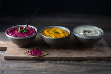 Three hummus dips, beet hummus, sweet potato or carrot or pumpkin hummus and parsnip hummus on a rustic wooden board with a purple napkin on a dark wooden table. Side view.