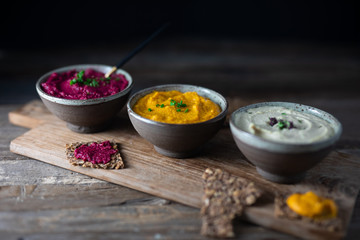 Three hummus dips, beet hummus, sweet potato or carrot or pumpkin hummus and parsnip hummus on a rustic wooden board with a purple napkin on a dark wooden table. Side view.