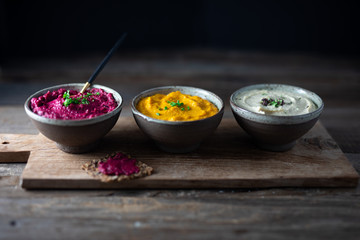Three hummus dips, beet hummus, sweet potato or carrot or pumpkin hummus and parsnip hummus on a rustic wooden board with a purple napkin on a dark wooden table. Side view.