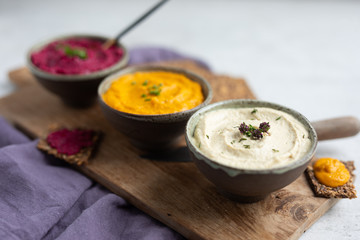 Three hummus dips, beet hummus, sweet potato or carrot or pumpkin hummus and parsnip hummus served with flax seeds crackers on a rustic wooden board with a spoon and a purple napkin on a light natural
