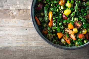 Du Puy lentils, cherry tomatoes, asparagus, sweet peas, scallions, parsley, cilantro, basil salad with vinaigrette in a pottery salad bowl. Top view, rustic wooden background, copy space. 