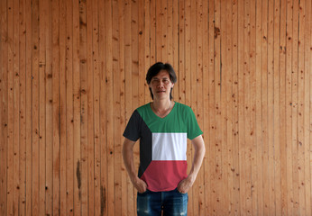 Man wearing Kuwait flag color shirt and standing with two hands in pant pockets on the wooden wall background.