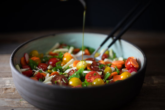 Fennel, flat green beans, cherry tomatoes, radish, carrot, cilantro, parsley, basil, hemp seeds, olive oil, balsamic vinegar salad in pottery dish with olive oil pouring. Side view