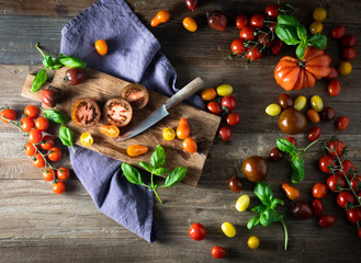 Top view of still life of orange, yellow, heirloom, dark red tomatoes and basil with a wooden chopping board, a rustic knife with a purple napkin. Top view, overhead, good for banner. 