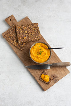 Sweet potato or pumpkin or carrot hummus with parsley and olive oil, served with seed crackers on a rustic wooden board with a spoon. Light background, top view, copy space.