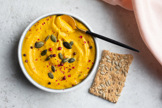 Top view of sweet potato or carrot or pumpkin hummus served with pink peppercorn and pumpkin seeds, a spoon and sunflower seeds crackers and a pink napkin. Flat lay, copy space. 