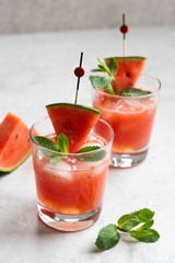 Two cold and fresh watermelon juices with ice cubes, fresh mint and a slice of watermelon on a light natural stone background. Side view, close up. 
