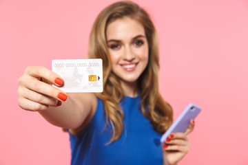 Beautiful happy young pretty woman posing isolated over pink wall background holding credit card using mobile phone.