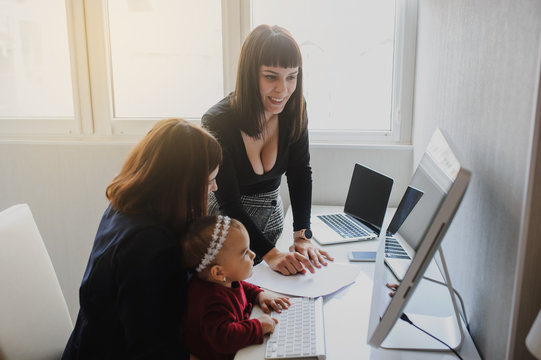 close up photo of two women in office outfit working on computers and discussing a report; one of them is with a baby girl in her arms