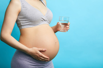 Young pregnant caucasian model on third trimester, dressed in grey underwear, holding a glass of water, isolated on light blue baclground with copyspace.