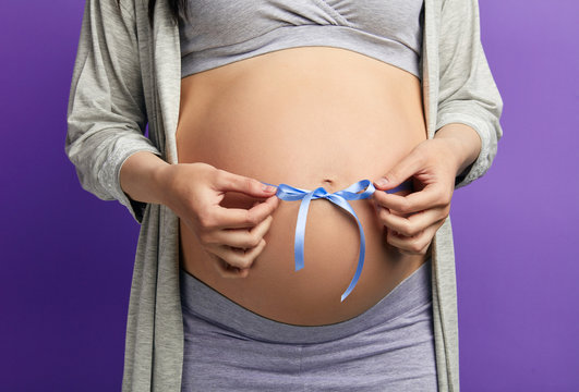 Cropped image of beautiful pregnant woman hugging her tummy with blue ribbon, expecting soon the precious gift- her newborn child.