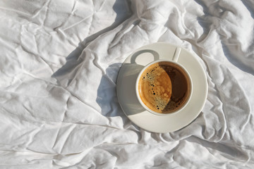 Cup of coffee on the bed with white linen in the room with a lot of natural sunlight, top view