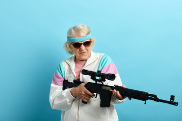 Do not make your grandmother angry. Grandma can respond. Comic portrait of old-aged grandma in whiye sportive outfit and dark sunglasses holding sniper rifle, pointing aside over blue background