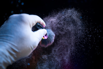 Naklejka na ściany i meble Makeup brush in hand with cosmetic powder on dark background with light and smoke. Powder splash on dark