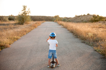 active sport little boy in helmet standing on empty asphalt road with scooter on summer warm sunset