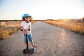 sport little caucasian kid in helmet and blue denim shorts yawn standing on empty asphalt road with scooter on summer warm sunset © ruslanshug