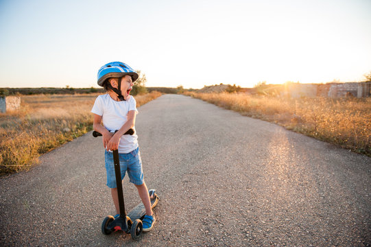 sport little caucasian kid in helmet and blue denim shorts yawn standing on empty asphalt road with scooter on summer warm sunset