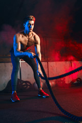 Achieving best results. Young male athlete in sports sweatpants doing cross fit workout with battle ropes on sportive show, performing on dark stage with red smoky background