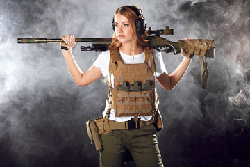 Skilled blonde female soldier with rifle in hands standing in military outfit in smoky darkness....