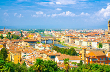 Top aerial panoramic view of Florence city with Ponte Vecchio bridge over Arno river, buildings with orange red tiled roofs, palm trees, blue sky white clouds, Tuscany, Italy