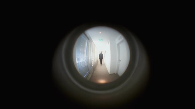 Seen from a spy hole point-of-view: a man in a suit, walking away from the door in a corridor.