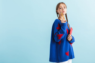 Young pretty girl with two braids in oversize sweater with red hearts holding notebook and pencil in hands thoughtfully looking in camera over blue background isolated