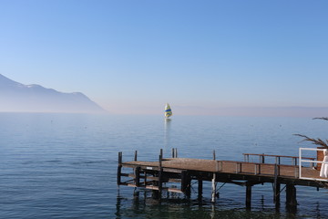 Small wooden dock stretching in to the lake with blue sky, mist abofe the lake and silhouette of mountain in background.