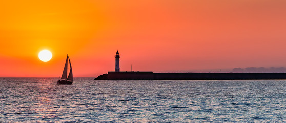 Panoramic view of ocean sunset with silhouettes of sailboat and lighthouse against the orange sky.