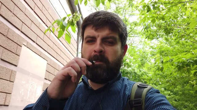 Bearded man in headphones is talking to the camera and smoking iqos against the background of green trees. Selfie.