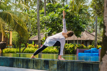 Beautiful young woman is practicing yoga on the edge of swimming pool with green plants and palm trees around. Sport photo with beautiful girl in luxury hotel on Bali island Indonesia